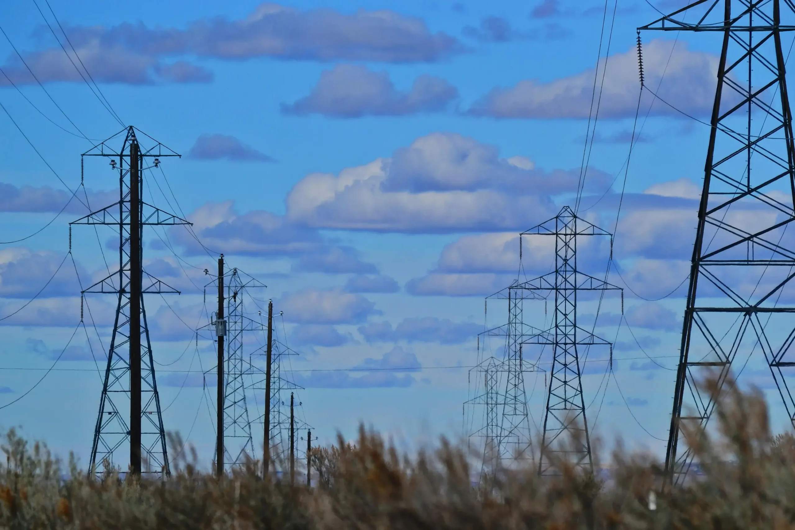 High-voltage transmission towers and power lines stretching across a rural landscape under a blue sky with scattered clouds