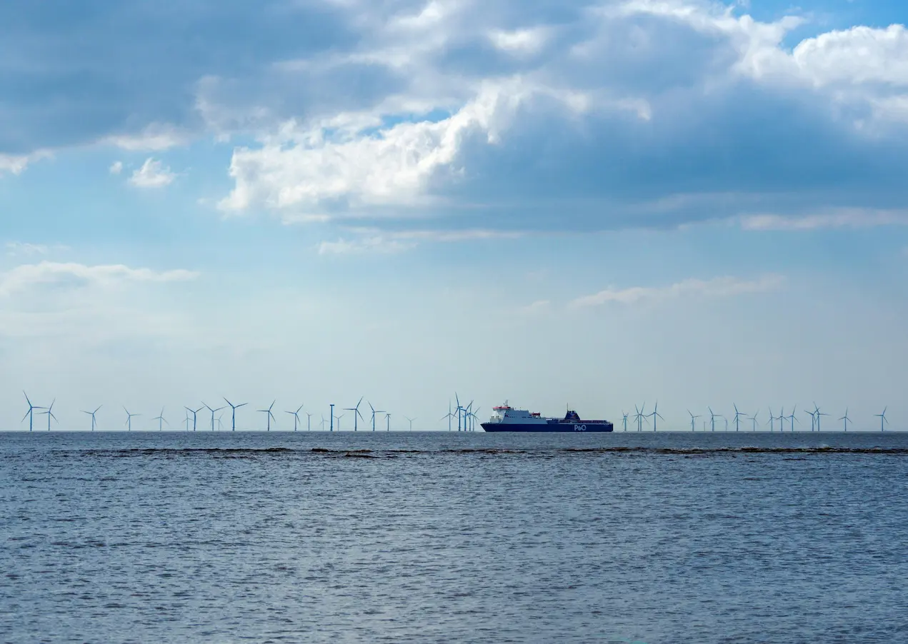 An offshore wind farm with rows of wind turbines on the horizon and a large cargo ferry sailing across the water under a cloudy sky